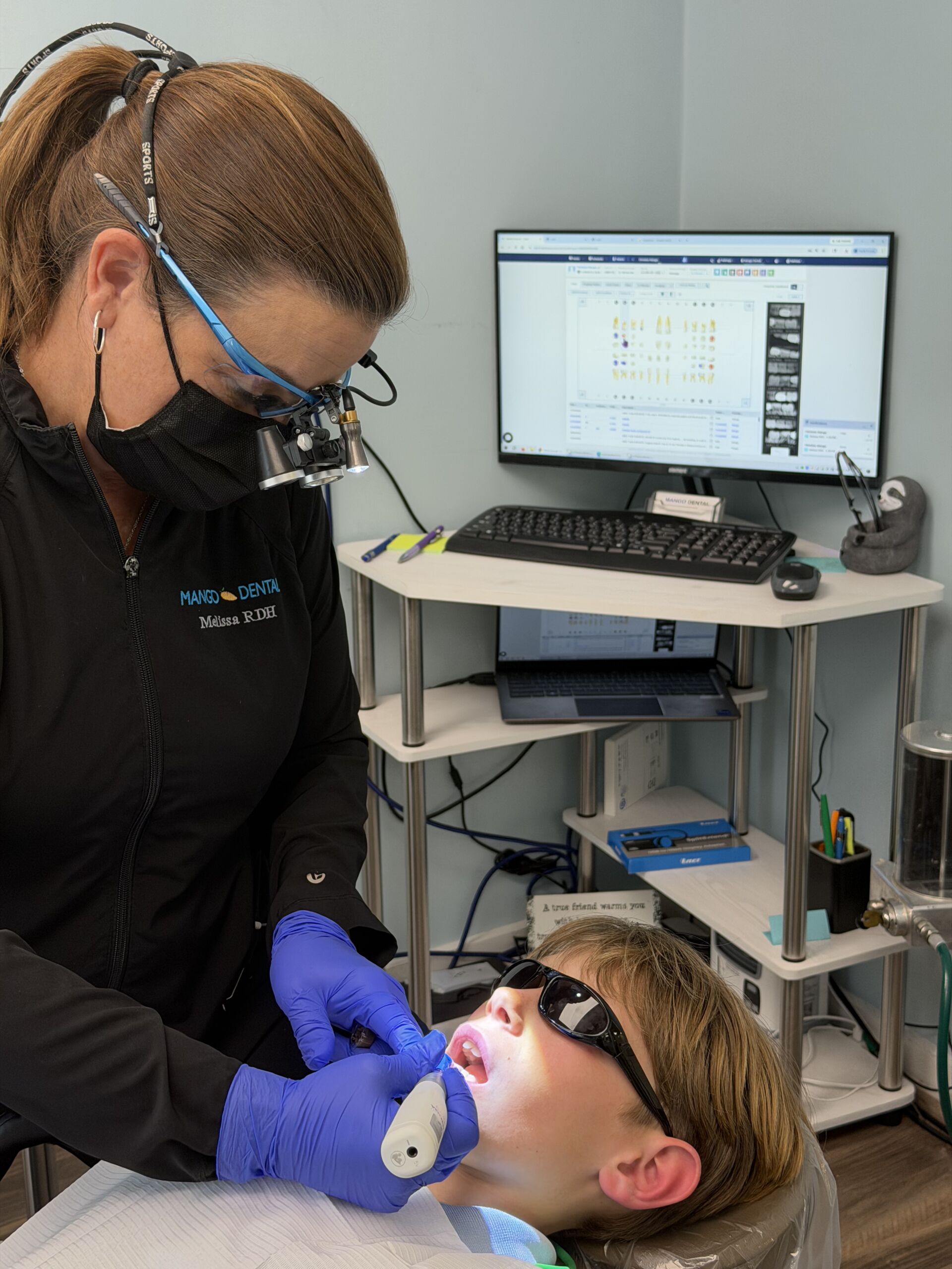 Dentist with magnifying loupes and blue gloves treats a pediatric patient in a dental chair.