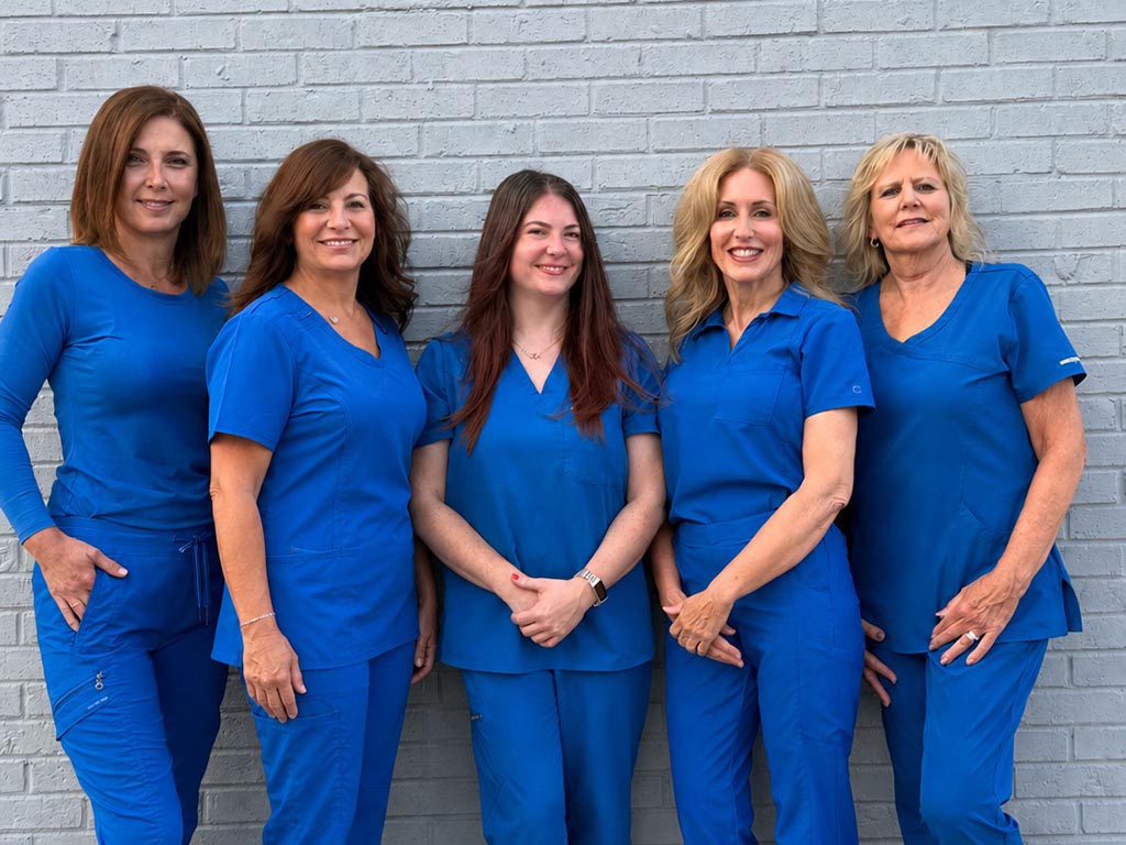 Mango Dental Hygiene Team - MANGO DENTAL Five women in blue scrubs posing together against a gray brick wall.